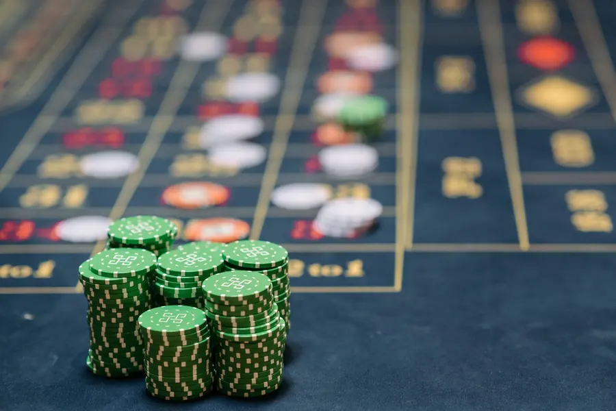 Stacks of green gaming chips in the foreground of a blue felt table layout, other chips scattered out of focus behind them