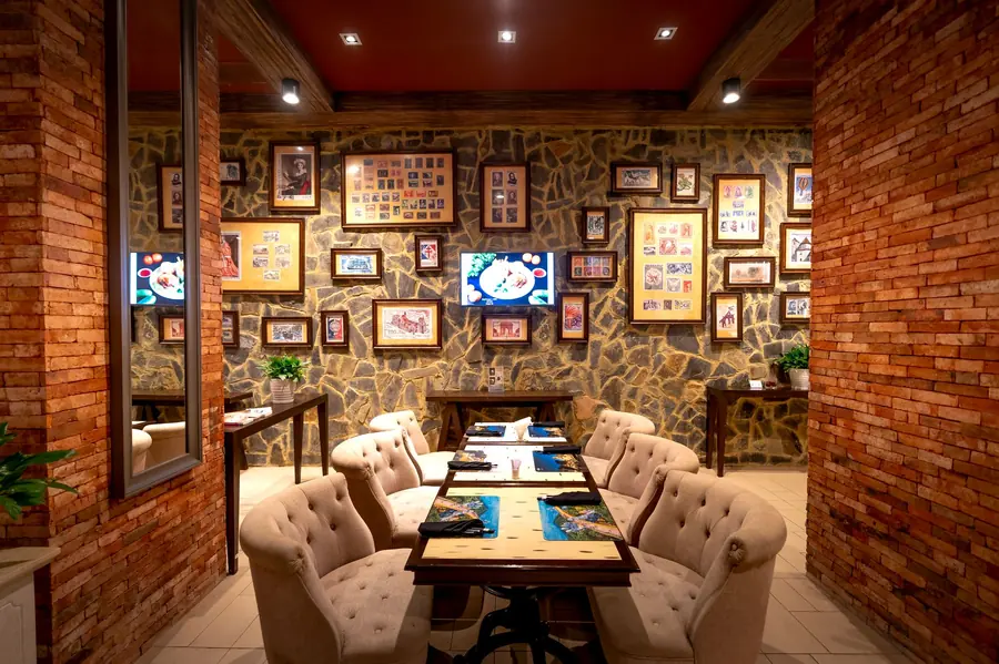 A narrow restaurant room with exposed brick and stone walls, framed pictures hung densely above a long table set with placemats and tub chairs