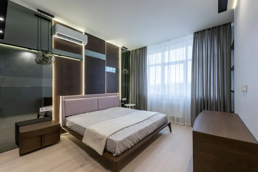 A modern hotel bedroom: low platform bed with a pale upholstered headboard, dark timber panelling, sheer curtains over a bright window