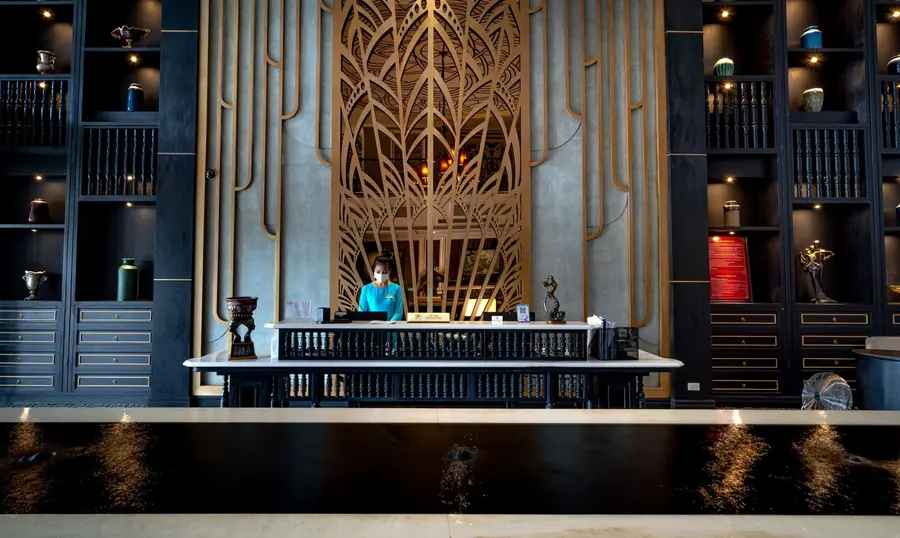 A hotel reception desk in a tall, dark-panelled lobby, an ornate gilt screen rising behind it and a member of staff at the counter