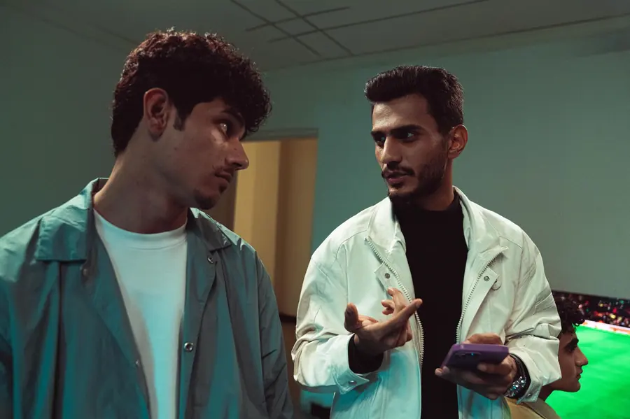 Two men standing indoors in conversation, one gesturing with an open hand while the other listens, both looking serious