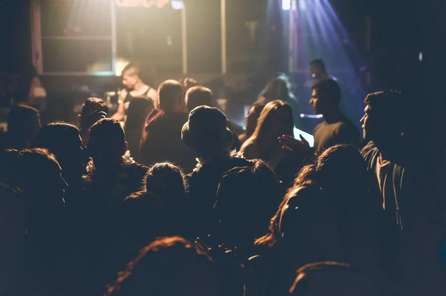 A concert audience photographed from behind in low light, heads silhouetted against warm stage lighting and a beam of blue light